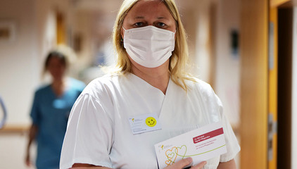 Eine Altenpflegekraft in weißer Funktionskleidung und Mundschutz blickt zum Internationalen Tag der Pflegenden mit einer Grußkarte in den Händen in die Kamera.