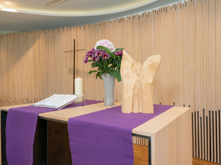 Kerze, Blumenstrauß und Holzengel auf Tisch im Raum der Stille