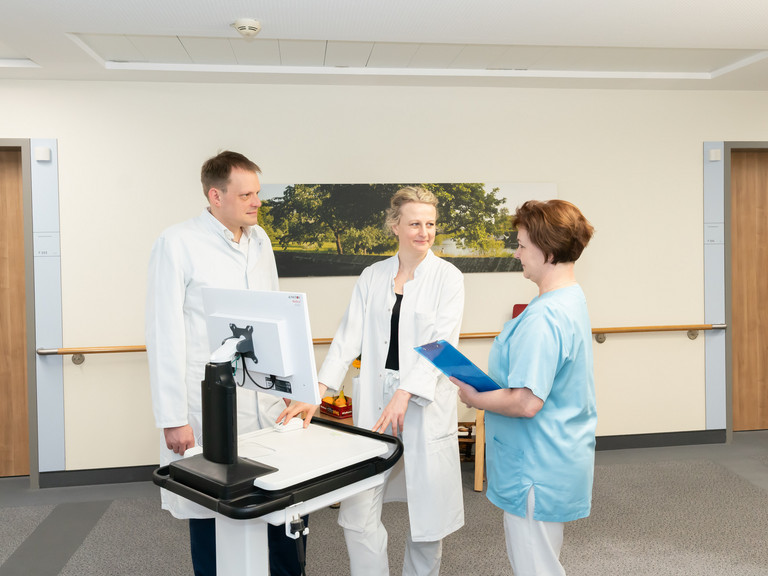 Arzt, Ärztin und Pflegfachfrau unterhalten sich auf Flur an einem mobilen Computerwagen - Immanuel Klinik Rüdersdorf - Neurologie - Neuromuskuläres Zentrum Arzt, Ärztin und Pflegfachfrau unterhalten sich auf Flur an einem mobilen Computerwagen - Immanuel Klinik Rüdersdorf - Neurologie - Neuromuskuläres Zentrum