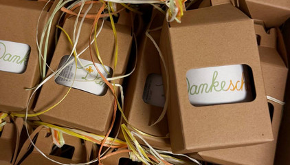 Mehrere in einer Kartonverpackung mit Sichtfenster verpackte weiße Tassen mit einer Aufschrift "Dankeschön" als grün-orangene Bändergrafik als Geschenk für Pflegekräfte am Internationalen Tag der Pflegenden.